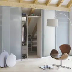 Przestronna garderoba pod niskim skosem, z drzwiami przesuwnymi, w której panuje porządek. Obok wygodny fotel i lampa.