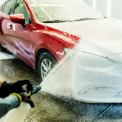 Czerwony samochód pokryty gęstą, białą pianą podczas mycia. Ręka w czarnej rękawiczce trzyma pistolet do piany, rozpylając ją na maskę pojazdu.