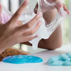 Dziecko bawi się przezroczystym klejem z mąki, który rozciąga się między palcami. Obok leżą dwie kupki niebieskiego slime'a.