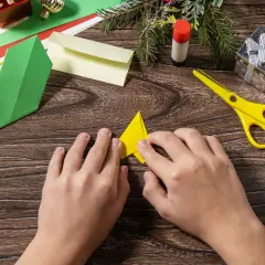 Dziecko składa żółty trójkąt, wybierając odpowiedni papier do origami. Obok leżą zielone, czerwone i żółte kartki, nożyczki i klej.