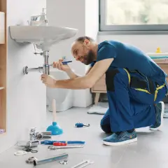 Hydraulik klęczy przy umywalce, używając klucza do pracy przy syfonie. Pokazuje, jak wymienić baterię w umywalce.