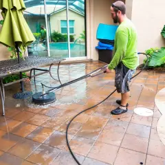 Mężczyzna czyści chropowaty gres na tarasie myjką ciśnieniową. Woda rozpryskuje się wokół.