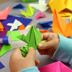 Dziecko tworzy zielonego żurawia z kartki A4. Wokół kolorowe origami, klej i papier. Z kartki papieru A4 można zrobić wiele pięknych rzeczy!