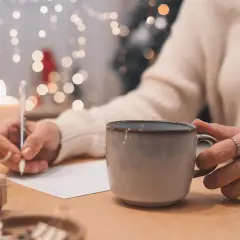 Osoba pisze podpis pod życzeniami urodzinowymi, trzymając kubek z gorącym napojem. W tle migoczą światełka choinkowe.