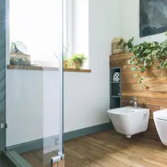 Nowoczesna łazienka z bidetem i miską WC. Drewniana ściana, rośliny i szklana kabina prysznicowa tworzą relaksującą atmosferę.