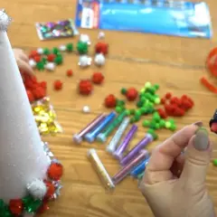 Ręce tworzą świąteczne ozdoby, używając pistoletu na klej i kolorowych pomponów. Wokół stołu leżą materiały do ozdób świątecznych.