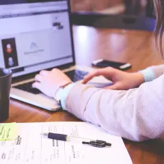 Kobieta pracuje przy laptopie. Na biurku leżą notatki, długopis i kubek. Idealne warunki pracy, odpowiednie ile luxów na stanowisku pracy.