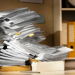 Stosy dokumentów czekają na archiwizację. Na biurku piętrzą się papiery, obok pudełko i przybory piśmiennicze.