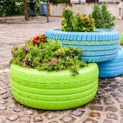 Kolorowe kwietniki z opon, wypełnione bujną zielenią i kwiatami, zdobią brukowany plac.
