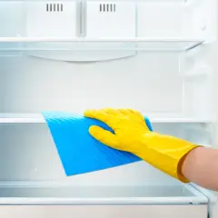 Ręka w żółtej rękawiczce czyści półkę lodówki niebieską ściereczką. Dowiedz się, czym myć lodówkę, by była lśniąca i higieniczna.