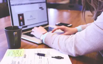 Kobieta pracuje przy laptopie. Na biurku leżą notatki, długopis i kubek. Idealne warunki pracy, odpowiednie ile luxów na stanowisku pracy.