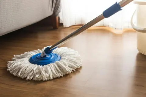 Mop z białymi sznurkami na niebieskiej podstawie, gotowy do czyszczenia paneli. Obok stoi wiadro.