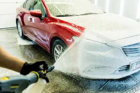 Czerwony samochód pokryty gęstą, białą pianą podczas mycia. Ręka w czarnej rękawiczce trzyma pistolet do piany, rozpylając ją na maskę pojazdu.