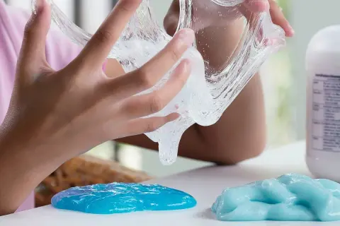 Dziecko bawi się przezroczystym klejem z mąki, który rozciąga się między palcami. Obok leżą dwie kupki niebieskiego slime'a.