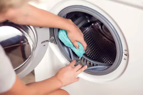 Ręce czyszczą uszczelkę pralki niebieską ściereczką. To prosty sposób, jak wyczyścić pralkę octem, usuwając kamień i zapachy.