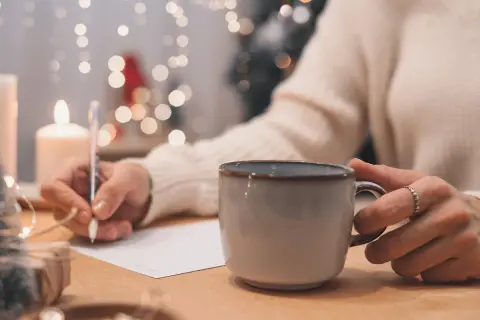 Osoba pisze podpis pod życzeniami urodzinowymi, trzymając kubek z gorącym napojem. W tle migoczą światełka choinkowe.