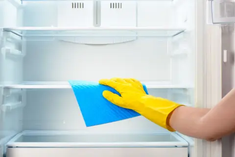 Ręka w żółtej rękawiczce czyści półkę lodówki niebieską ściereczką. Dowiedz się, czym myć lodówkę, by była lśniąca i higieniczna.