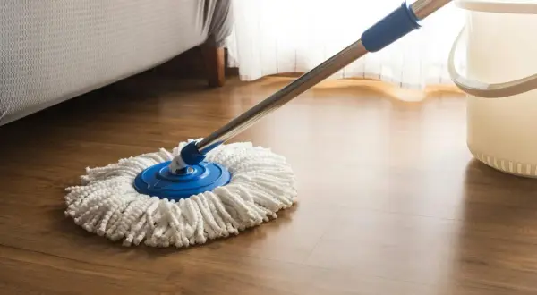 Mop z białymi sznurkami na niebieskiej podstawie, gotowy do czyszczenia paneli. Obok stoi wiadro.