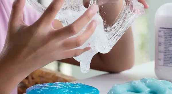 Dziecko bawi się przezroczystym klejem z mąki, który rozciąga się między palcami. Obok leżą dwie kupki niebieskiego slime'a.