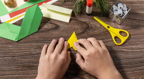 Dziecko składa żółty trójkąt, wybierając odpowiedni papier do origami. Obok leżą zielone, czerwone i żółte kartki, nożyczki i klej.