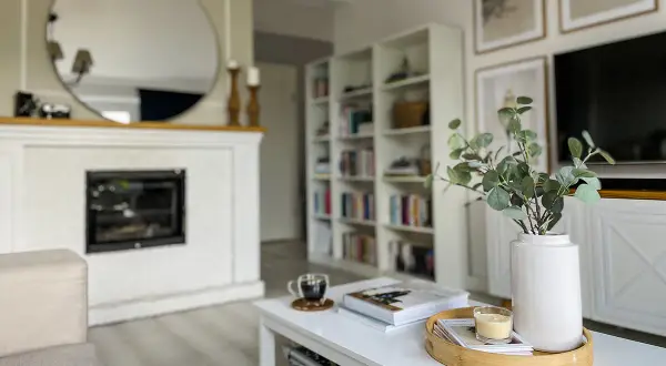 Biały stolik kawowy DIY z książkami, świecą i wazonem z gałązkami eukaliptusa. W tle kominek i regał z książkami.