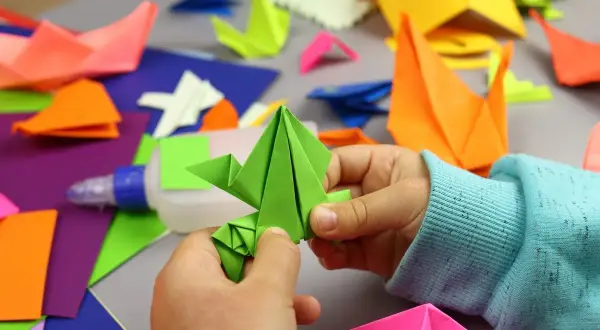 Dziecko tworzy zielonego żurawia z kartki A4. Wokół kolorowe origami, klej i papier. Z kartki papieru A4 można zrobić wiele pięknych rzeczy!