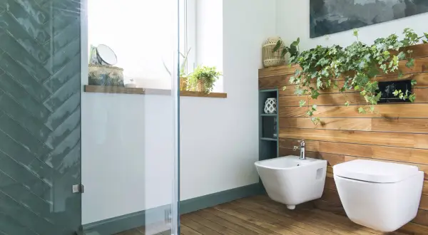 Nowoczesna łazienka z bidetem i miską WC. Drewniana ściana, rośliny i szklana kabina prysznicowa tworzą relaksującą atmosferę.