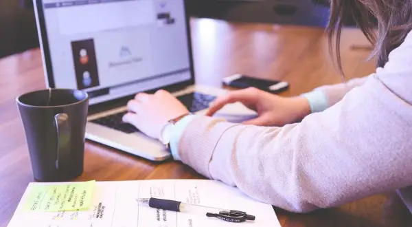 Kobieta pracuje przy laptopie. Na biurku leżą notatki, długopis i kubek. Idealne warunki pracy, odpowiednie ile luxów na stanowisku pracy.