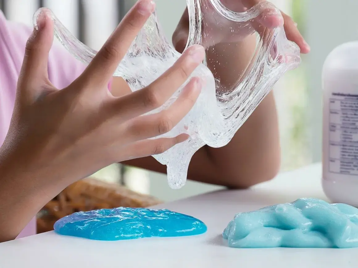Dziecko bawi się przezroczystym klejem z mąki, który rozciąga się między palcami. Obok leżą dwie kupki niebieskiego slime'a.