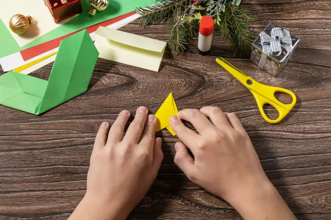 Dziecko składa żółty trójkąt, wybierając odpowiedni papier do origami. Obok leżą zielone, czerwone i żółte kartki, nożyczki i klej.