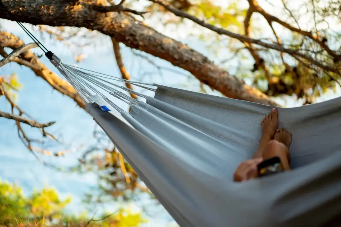 Relaks w hamaku z drążkiem czy bez? Stopy odpoczywają, telefon w dłoni, drzewa wokół.