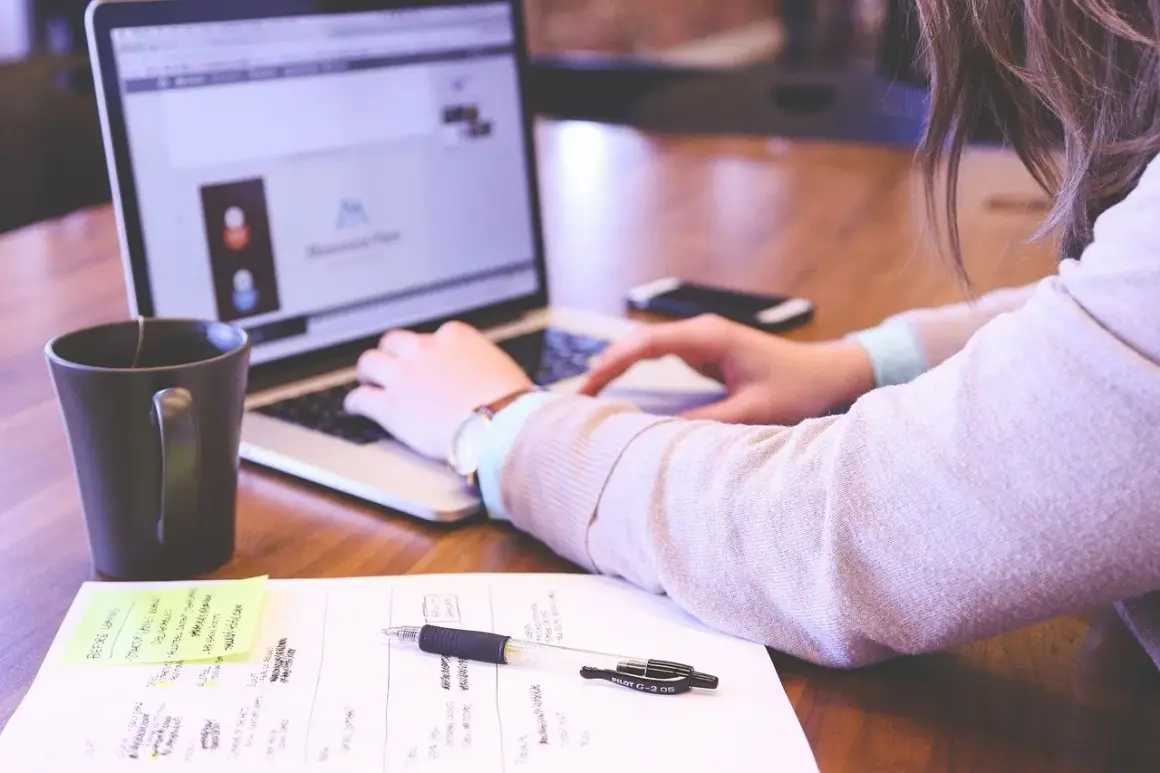 Kobieta pracuje przy laptopie. Na biurku leżą notatki, długopis i kubek. Idealne warunki pracy, odpowiednie ile luxów na stanowisku pracy.
