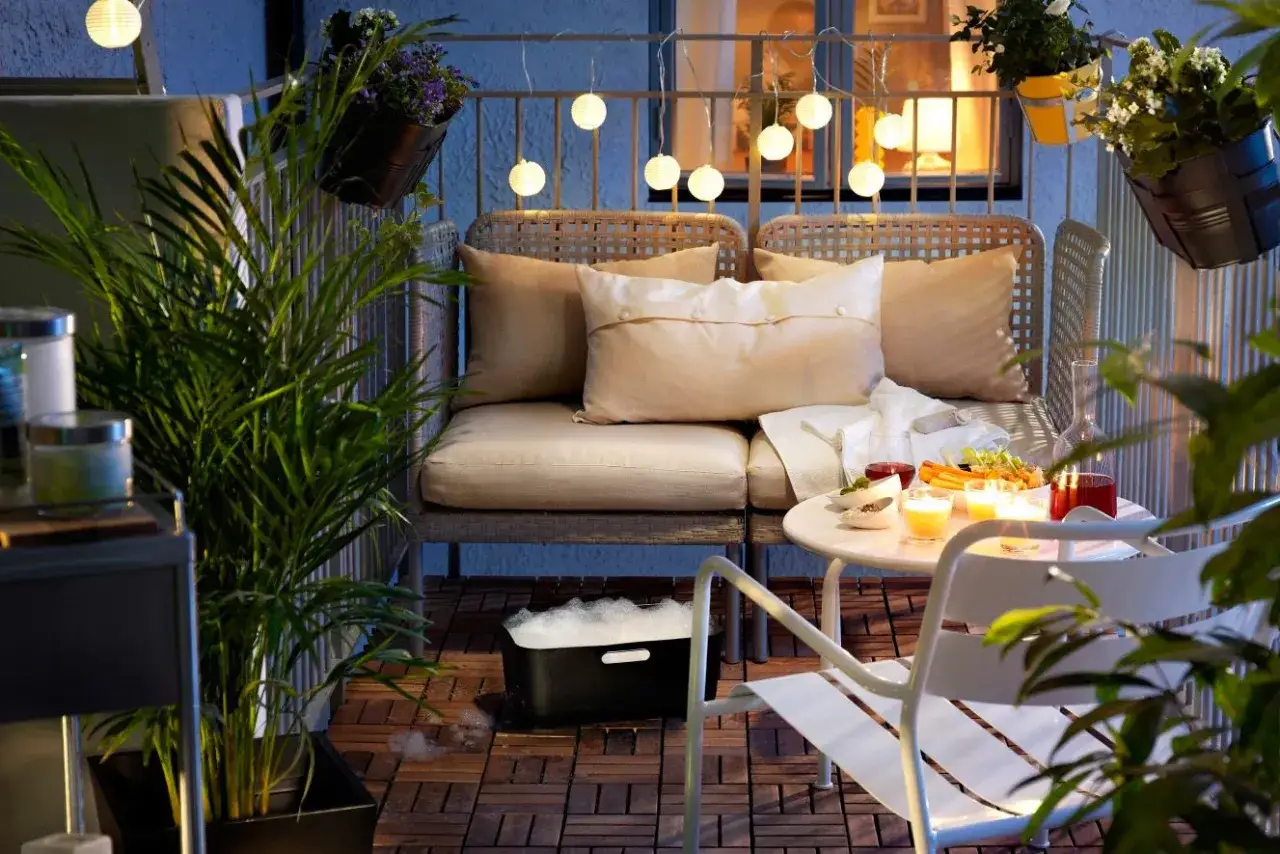Przytulny balkon w bloku z wygodną sofą, stolikiem z przekąskami i świecami. Girlanda świetlna dodaje nastroju.