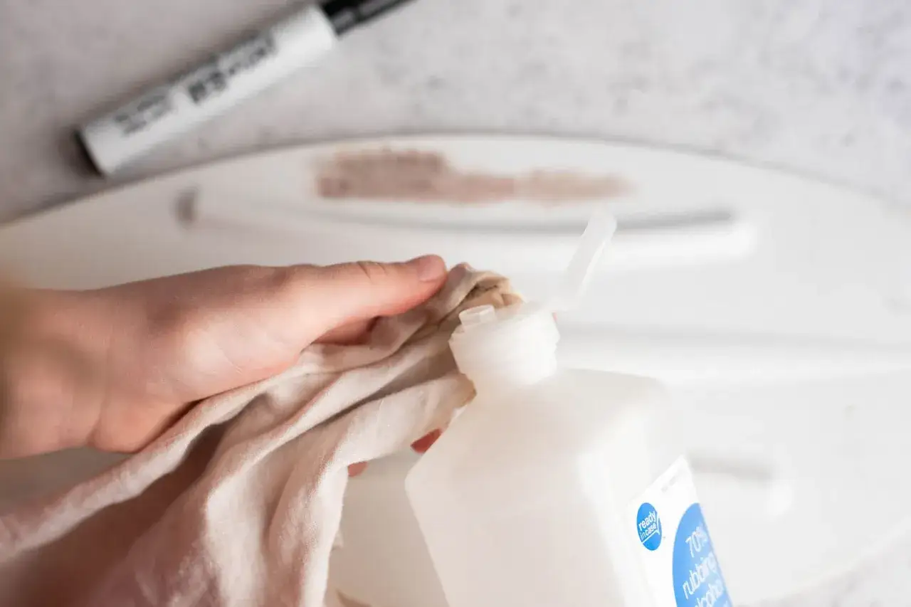 Ręka z szmatką nasączoną alkoholem próbuje zmyć ślady markera z białego plastiku. Dowiedz się, czym zmyć marker z plastiku.