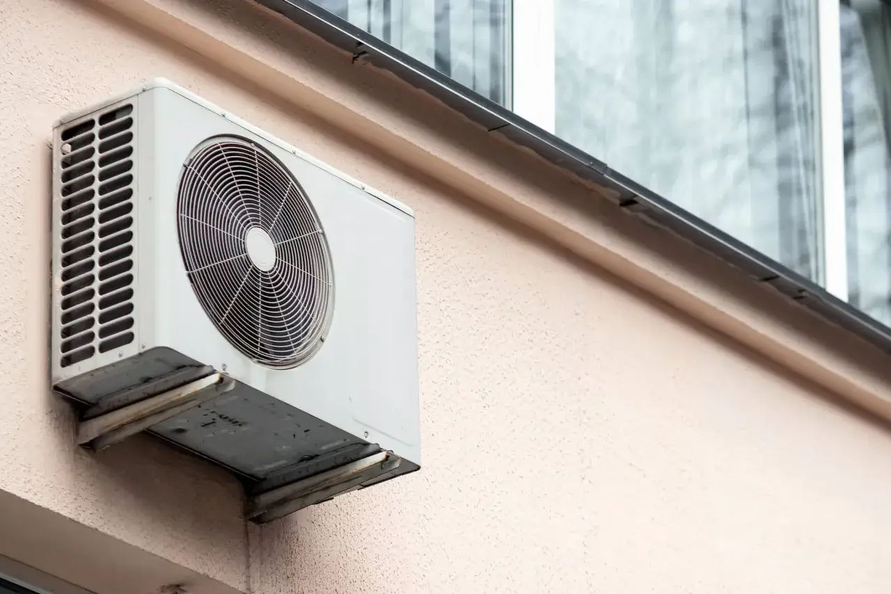 Biały klimatyzator zamontowany na ścianie budynku. Widać jego wentylator i kratkę. Obok okno sąsiada.