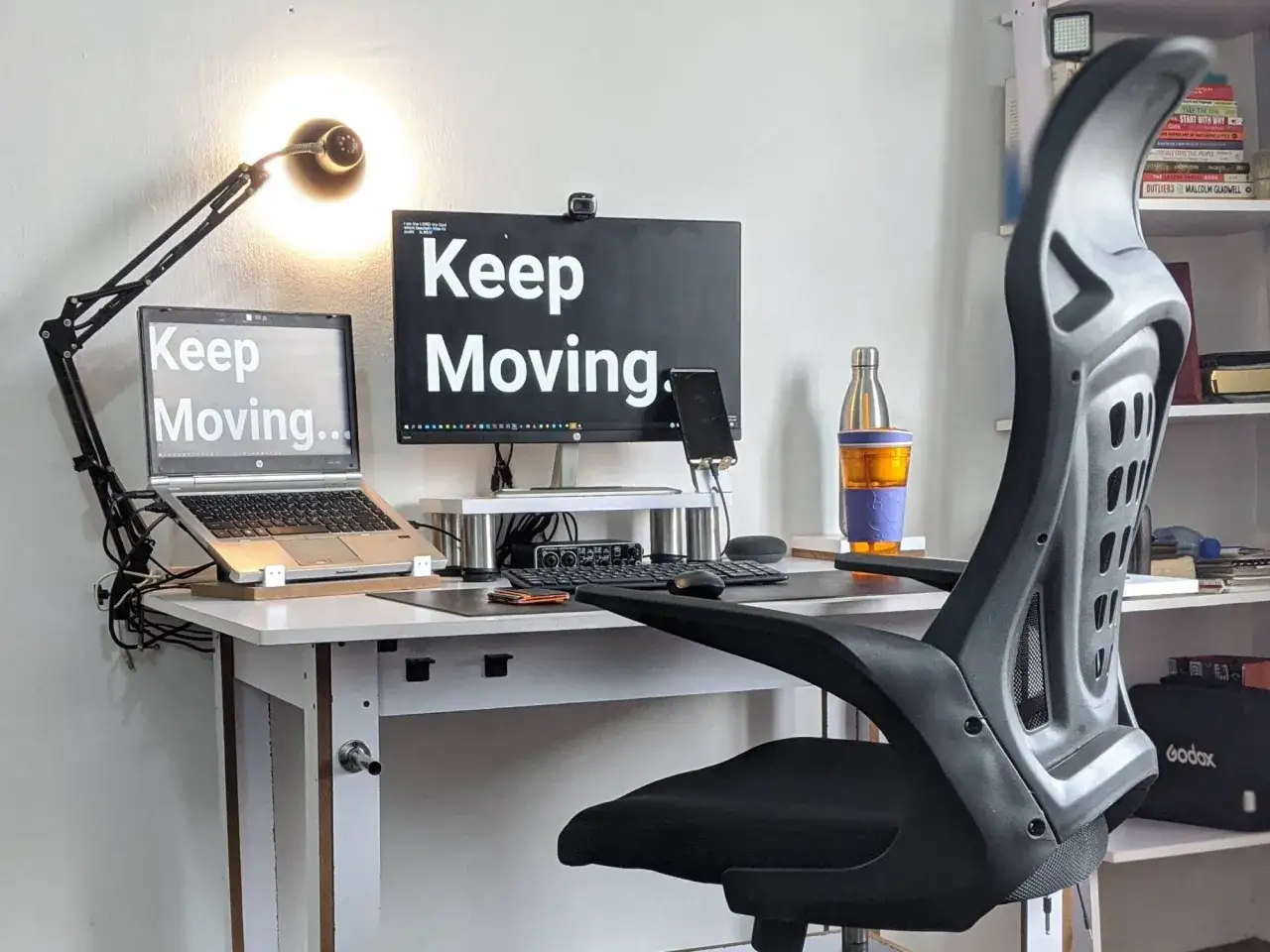 Jak urządzić biuro w domu? Ergonomiczne krzesło, biurko z monitorem i laptopem, lampka i motywujące napisy