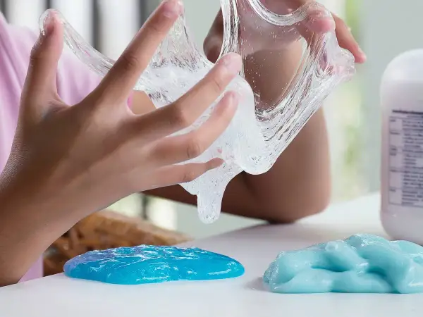 Dziecko bawi się przezroczystym klejem z mąki, który rozciąga się między palcami. Obok leżą dwie kupki niebieskiego slime'a.