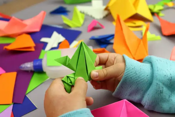 Dziecko tworzy zielonego żurawia z kartki A4. Wokół kolorowe origami, klej i papier. Z kartki papieru A4 można zrobić wiele pięknych rzeczy!