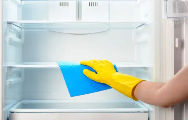 Ręka w żółtej rękawiczce czyści półkę lodówki niebieską ściereczką. Dowiedz się, czym myć lodówkę, by była lśniąca i higieniczna.